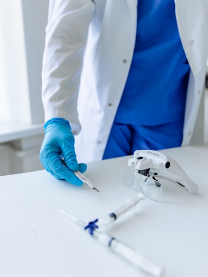 A medical professional wearing a white coat and blue scrubs is handling a scalpel. The individual is also wearing blue gloves. On the table in front are medical instruments, including syringes and a device possibly used for magnification purposes, placed on a clean white surface.