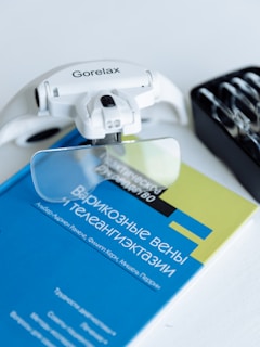 A white device labeled 'Gorelax' with optical lenses is placed on top of a blue and yellow book with Cyrillic text. The book appears to discuss varicose veins. A black tray with various tools or lenses is partially visible on the right side.