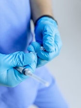 A person wearing blue medical scrubs and gloves is handling a syringe, potentially preparing it for use. The focus is on the hands and syringe, indicating a medical or healthcare setting.
