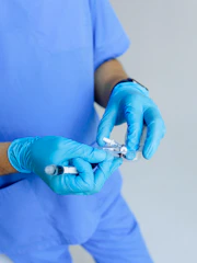 A person wearing a blue medical uniform and blue gloves is holding a syringe and a vial, likely preparing an injection.