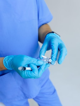 A person wearing a blue medical uniform and blue gloves is holding a syringe and a vial, likely preparing an injection.