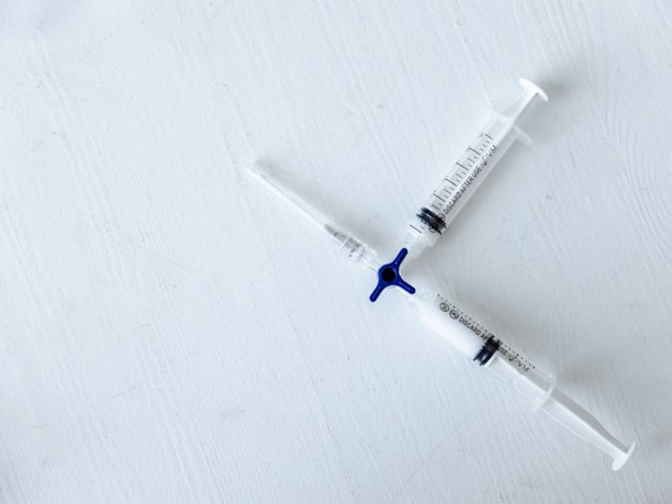 Two syringes intersect at right angles on a white textured surface. Each syringe has clear measurements visible and features a blue valve at the intersection point.