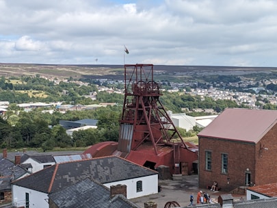A scenic view of a historical mining site featuring a prominent red mining tower surrounded by various industrial buildings. The background presents a lush landscape with dense greenery and a town visible in the distance. A flag can be seen atop the tower, and a few people are gathered near the buildings, adding a touch of activity to the scene.