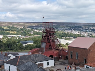 A scenic view of a historical mining site featuring a prominent red mining tower surrounded by various industrial buildings. The background presents a lush landscape with dense greenery and a town visible in the distance. A flag can be seen atop the tower, and a few people are gathered near the buildings, adding a touch of activity to the scene.