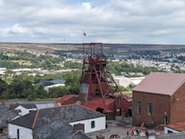 A scenic view of a historical mining site featuring a prominent red mining tower surrounded by various industrial buildings. The background presents a lush landscape with dense greenery and a town visible in the distance. A flag can be seen atop the tower, and a few people are gathered near the buildings, adding a touch of activity to the scene.