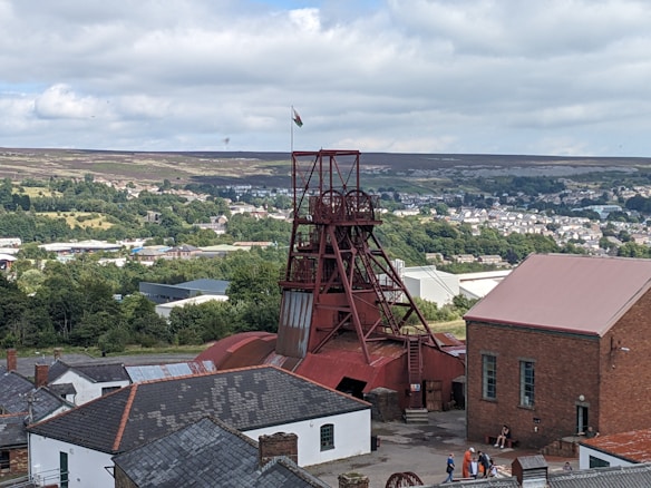 A scenic view of a historical mining site featuring a prominent red mining tower surrounded by various industrial buildings. The background presents a lush landscape with dense greenery and a town visible in the distance. A flag can be seen atop the tower, and a few people are gathered near the buildings, adding a touch of activity to the scene.