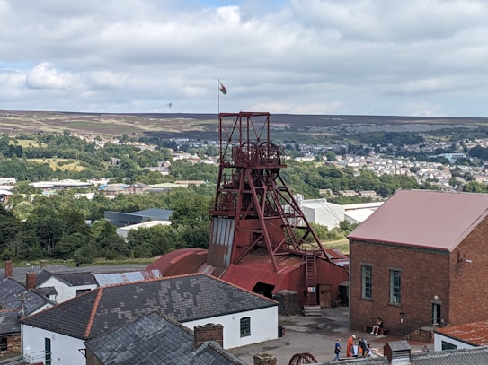 A scenic view of a historical mining site featuring a prominent red mining tower surrounded by various industrial buildings. The background presents a lush landscape with dense greenery and a town visible in the distance. A flag can be seen atop the tower, and a few people are gathered near the buildings, adding a touch of activity to the scene.