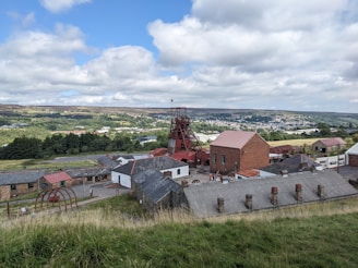A historical mining site with a large red metal tower structure, surrounded by several brick and stone buildings with slate roofs. The landscape in the background features rolling hills and stretches of grass and trees, with a town visible in the distance under a partly cloudy sky.