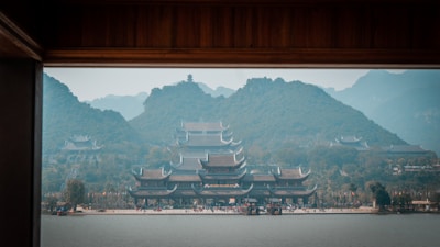 A traditional Asian temple with intricate roofs surrounded by lush green mountains. The scene includes a calm body of water in the foreground and misty hills in the background, creating a serene and picturesque landscape.