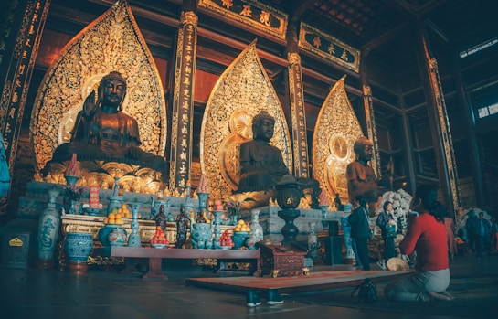 A richly decorated Buddhist temple interior features three large Buddha statues on an ornate altar. The statues are surrounded by detailed golden backdrops and adorned with various offerings, including fruit and vases. Visitors are seen kneeling and observing the serene setting.
