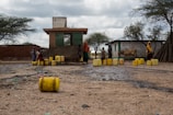 Several individuals, including children, are gathered around a small building with multiple yellow jerry cans scattered on the ground. The scene appears to be in a rural area with trees and a few structures nearby. A donkey is also present, tethered next to the people.