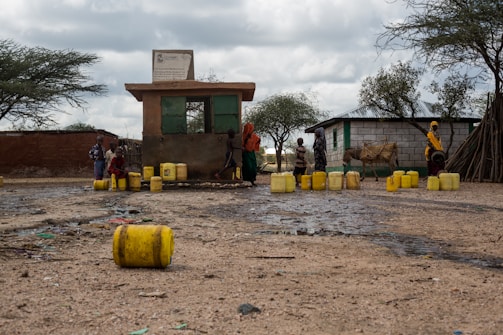 Several individuals, including children, are gathered around a small building with multiple yellow jerry cans scattered on the ground. The scene appears to be in a rural area with trees and a few structures nearby. A donkey is also present, tethered next to the people.