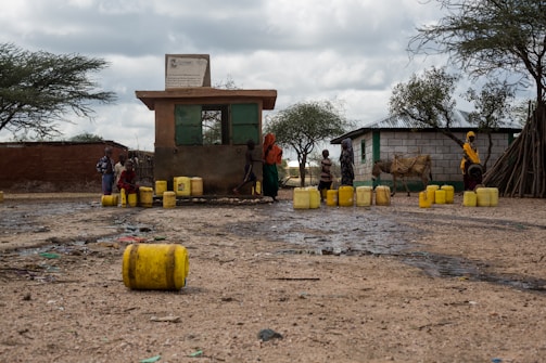 Several individuals, including children, are gathered around a small building with multiple yellow jerry cans scattered on the ground. The scene appears to be in a rural area with trees and a few structures nearby. A donkey is also present, tethered next to the people.
