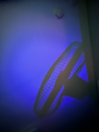 A ceiling fan with a protective grille is casting a shadow against a wall. The room is dimly lit with a blue hue, creating a calming and moody atmosphere.
