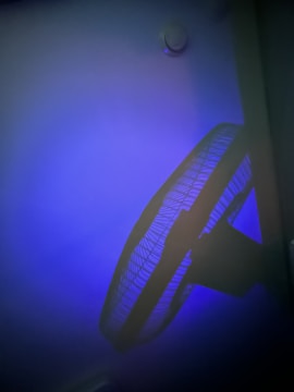 A ceiling fan with a protective grille is casting a shadow against a wall. The room is dimly lit with a blue hue, creating a calming and moody atmosphere.