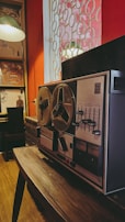 A vintage reel-to-reel tape recorder resting on a wooden table in a dimly lit room with red walls. The room features a window with circular patterned bars and a bookshelf filled with various items, including a large painting of an expressive face. A table lamp with a pleated shade hangs on the left side.