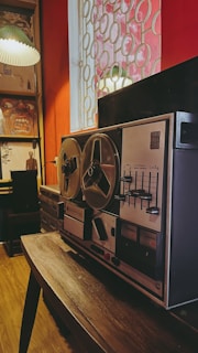 A vintage reel-to-reel tape recorder resting on a wooden table in a dimly lit room with red walls. The room features a window with circular patterned bars and a bookshelf filled with various items, including a large painting of an expressive face. A table lamp with a pleated shade hangs on the left side.