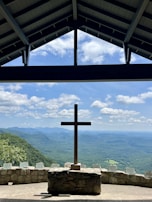 Under a shelter with a triangular roof, a large wooden cross stands prominently against a backdrop of rolling hills and distant mountains under a partly cloudy blue sky. The landscape is lush and green, with a stone wall enclosing the area and several plaques or markers placed along it.