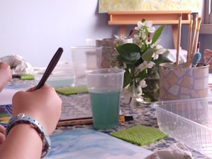A person is drawing with a pencil or pen on white paper, surrounded by art supplies including a palette, watercolor paints, paintbrushes, and a cup of water. White flowers and greenery are placed nearby, adding a natural element to the scene. The atmosphere is calm and creative.