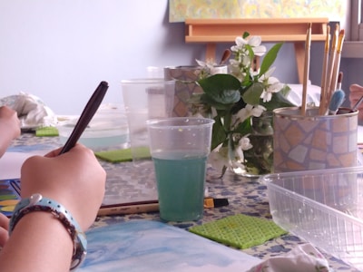 A person is drawing with a pencil or pen on white paper, surrounded by art supplies including a palette, watercolor paints, paintbrushes, and a cup of water. White flowers and greenery are placed nearby, adding a natural element to the scene. The atmosphere is calm and creative.