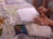 A person is painting with watercolors on a piece of paper. Various painting supplies are visible, including a palette with multiple watercolor paints, paintbrushes, and cups filled with water. The table is covered with a floral-patterned cloth.