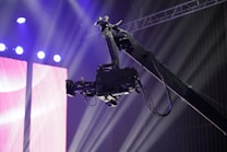 A professional video camera is mounted on a crane or jib arm against a backdrop of bright stage lights and colorful screens. The setup suggests a live event or production setting, with beams of light shining through the space.