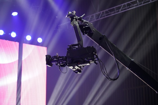 A professional video camera is mounted on a crane or jib arm against a backdrop of bright stage lights and colorful screens. The setup suggests a live event or production setting, with beams of light shining through the space.