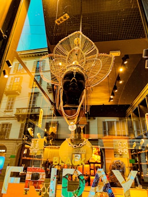 A store display features an elaborate mannequin headpiece adorned with intricate golden and metallic designs. The headpiece has multiple layers, with spokes and points extending outward. The background shows a reflection of a city street, with buildings visible through the glass. Bright lights inside the store illuminate the headpiece, creating a dramatic contrast. 'BLACK FRIDAY' is displayed in colorful letters at the bottom.
