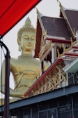 A large golden Buddha statue stands beside an intricately decorated building with traditional Thai architectural elements, including ornate patterns and pointed roofs. The structure features red and gold detailing, with the statue's serene expression gazing outward. A portion of a red awning or structure partially frames the left side of the image.