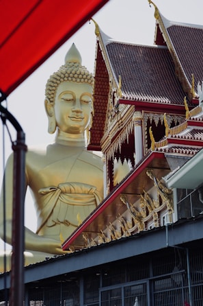 A large golden Buddha statue stands beside an intricately decorated building with traditional Thai architectural elements, including ornate patterns and pointed roofs. The structure features red and gold detailing, with the statue's serene expression gazing outward. A portion of a red awning or structure partially frames the left side of the image.
