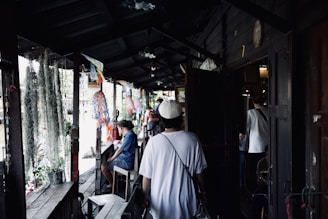A narrow wooden walkway with a group of people casually moving and sitting around. Decorations hang from the ceiling, and there are plants placed along the sides. The setting appears relaxed, possibly in a market or communal area.