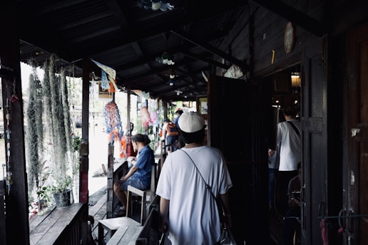 A narrow wooden walkway with a group of people casually moving and sitting around. Decorations hang from the ceiling, and there are plants placed along the sides. The setting appears relaxed, possibly in a market or communal area.