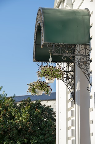 A green awning with ornate metalwork is attached to a building, beneath which two hanging baskets filled with small purple and white flowers are visible. Below, lush greenery and a partial view of another building can be seen against a clear blue sky.