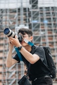 A person wearing a black shirt and watch is holding a professional camera with a large lens, standing against a background of scaffolding or a construction site. The person is focused on capturing a photograph and is equipped with a backpack and camera strap.