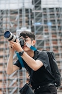 A person wearing a black shirt and watch is holding a professional camera with a large lens, standing against a background of scaffolding or a construction site. The person is focused on capturing a photograph and is equipped with a backpack and camera strap.