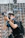 A person wearing a black shirt and watch is holding a professional camera with a large lens, standing against a background of scaffolding or a construction site. The person is focused on capturing a photograph and is equipped with a backpack and camera strap.