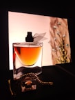 A stylish perfume bottle with a black cap stands elegantly in front of a softly lit background. The liquid inside the bottle appears amber-colored. In the background, delicate blue flowers adorn a fabric-like texture, adding a touch of nature and sophistication. A gold chain accessory is casually draped around the bottle, enhancing the luxurious aesthetic.