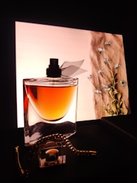 A stylish perfume bottle with a black cap stands elegantly in front of a softly lit background. The liquid inside the bottle appears amber-colored. In the background, delicate blue flowers adorn a fabric-like texture, adding a touch of nature and sophistication. A gold chain accessory is casually draped around the bottle, enhancing the luxurious aesthetic.