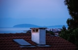 A serene rooftop scene featuring a red-tiled roof with a prominent chimney in the foreground. A skylight can be seen on the roof, and lush green foliage is visible on the right side. In the background, there's a calm expanse of water with a distant landmass, under a soft, dusky sky suggesting either dawn or dusk.