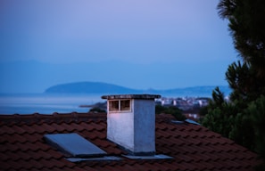 A serene rooftop scene featuring a red-tiled roof with a prominent chimney in the foreground. A skylight can be seen on the roof, and lush green foliage is visible on the right side. In the background, there's a calm expanse of water with a distant landmass, under a soft, dusky sky suggesting either dawn or dusk.
