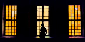 A silhouette of a person stands in front of three large rectangular windows with yellow and orange tinted glass panes. The windows are evenly spaced, and the contrast between the dark interior and the brightly colored exterior creates a striking visual effect.
