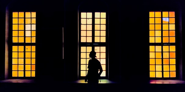 A silhouette of a person stands in front of three large rectangular windows with yellow and orange tinted glass panes. The windows are evenly spaced, and the contrast between the dark interior and the brightly colored exterior creates a striking visual effect.