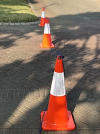 A series of bright orange traffic cones are positioned in a line on a brick-paved surface. A blue rope is connected between the cones, casting shadows on the ground due to sunlight. In the background, there is a grassy area and a curved path.