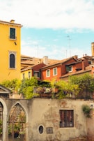 A charming scene of colorful Italian houses with terracotta roofs, featuring a vibrant yellow and a deep red facade. Lush greenery adorns the terrace of a house, adding a touch of nature. An old stone wall with a rustic metal gate and a barred window can be seen in the foreground, enhancing the historical ambiance of the setting.