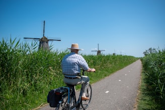 An elderly person wearing a hat is cycling on a path surrounded by tall green grass. The sky is clear and blue, and in the background, several traditional windmills are visible.