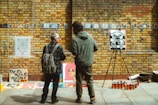 Two people stand in front of a brick wall displaying various artworks. The wall is adorned with small framed images and a large QR code. An easel holds an abstract black-and-white painting, and several colorful spray paint cans are lined up on the ground. A sign reads 'Pay as much as you can 4 art'. One person has a backpack and appears to be discussing the art with the other.