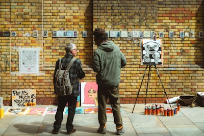 Two people stand in front of a brick wall displaying various artworks. The wall is adorned with small framed images and a large QR code. An easel holds an abstract black-and-white painting, and several colorful spray paint cans are lined up on the ground. A sign reads 'Pay as much as you can 4 art'. One person has a backpack and appears to be discussing the art with the other.