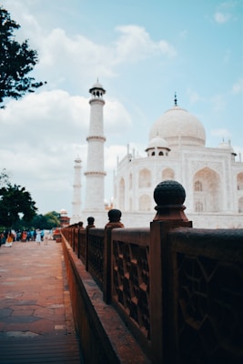 A well-known marble mausoleum with intricate designs stands prominently, characterized by its large dome and tall minarets. A decorative stone railing lines the foreground, leading towards the structure. People are visible walking towards the building, and trees flank the scene under a partly cloudy sky.