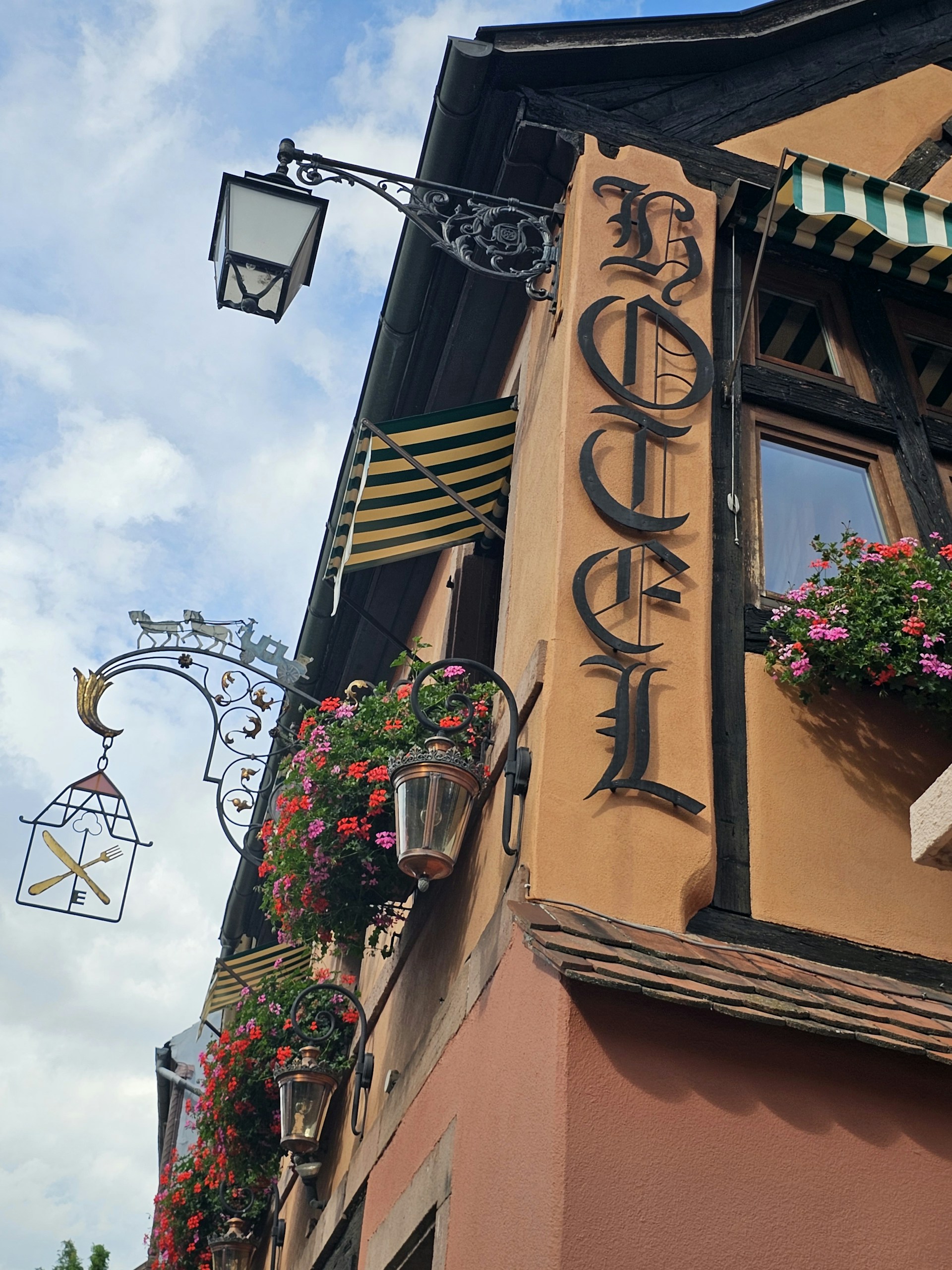 A vintage hotel facade features ornate wrought iron light fixtures and hanging flower baskets. The sign reads 'Hotel' in Gothic lettering, and striped awnings add to the charming, old-world aesthetic.