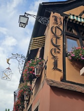 A vintage hotel facade features ornate wrought iron light fixtures and hanging flower baskets. The sign reads 'Hotel' in Gothic lettering, and striped awnings add to the charming, old-world aesthetic.